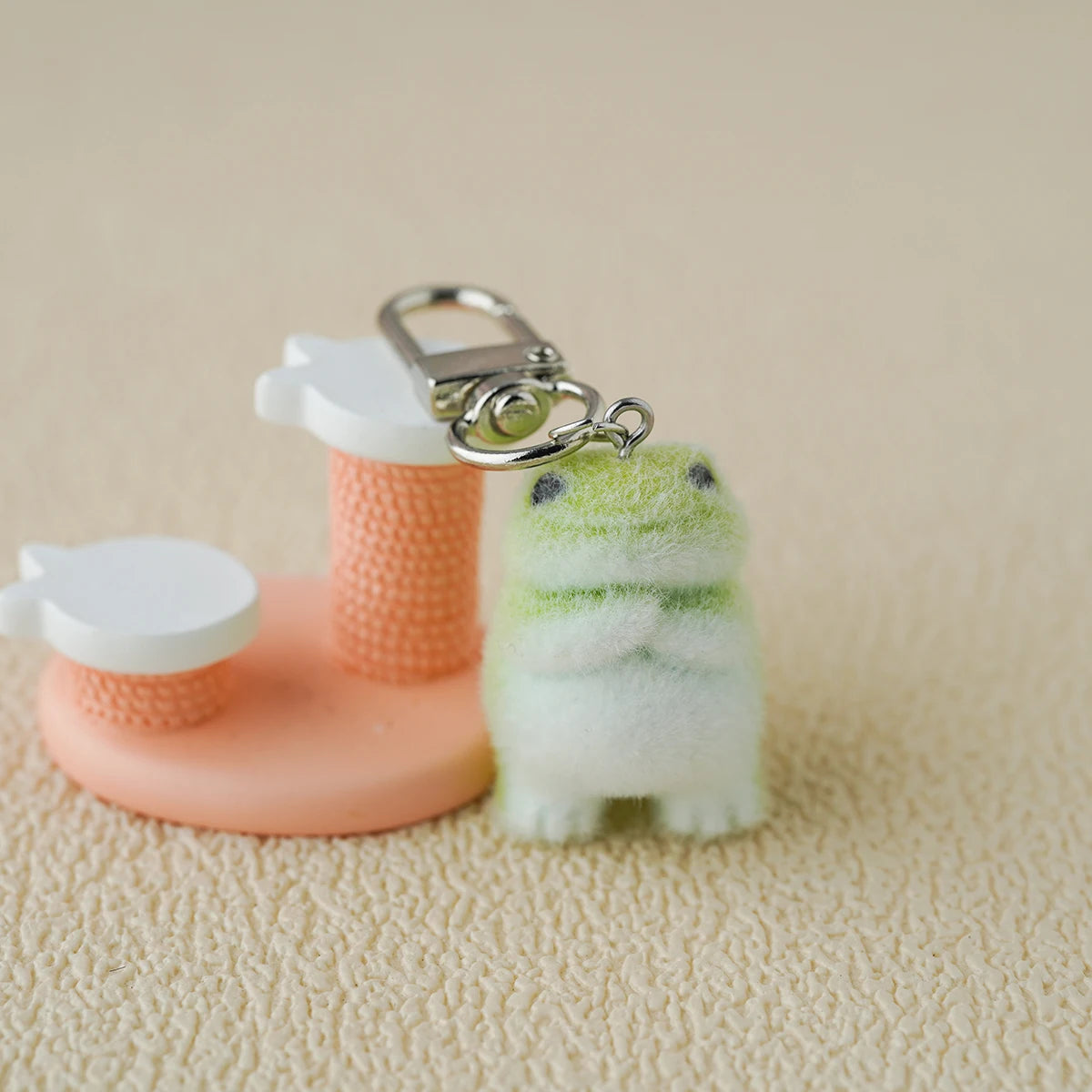 A tiny plush emotional‑support frog keychain with soft green fuzz, standing with his arms crossed beside a pink mini stand like a little guy waiting to be adopted