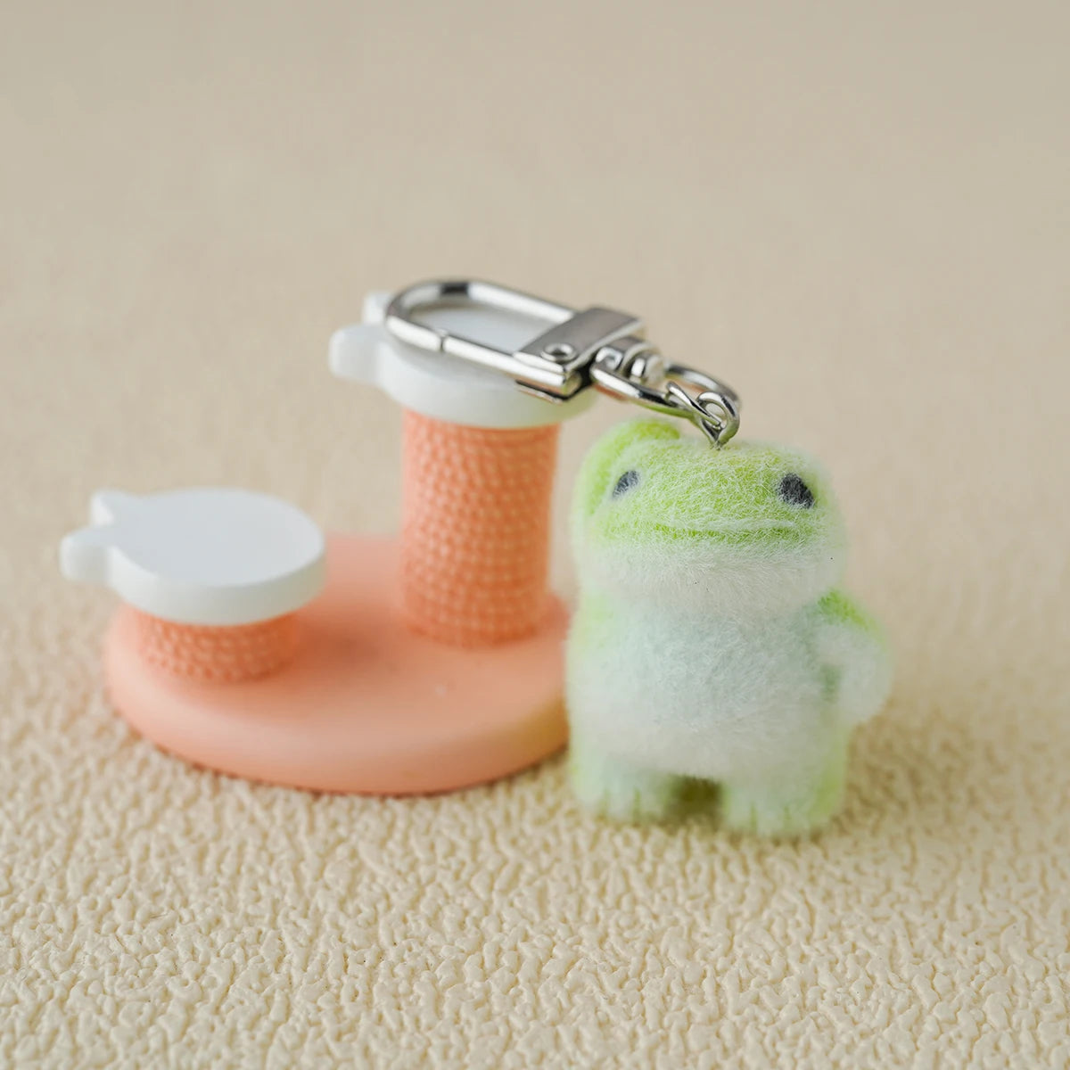 A tiny plush emotional‑support frog keychain with soft green fuzz, hanging beside a pink mini stand like a little guy waiting to be adopted