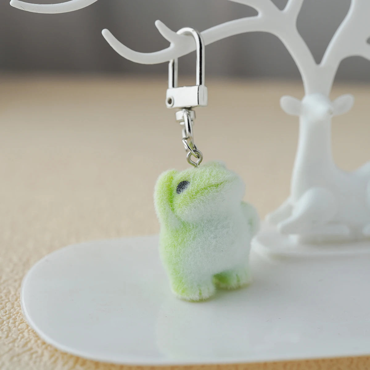 A tiny plush emotional‑support frog keychain with soft green fuzz, dangling from a metal clasp on a white stand like a little guy awaiting adoption
