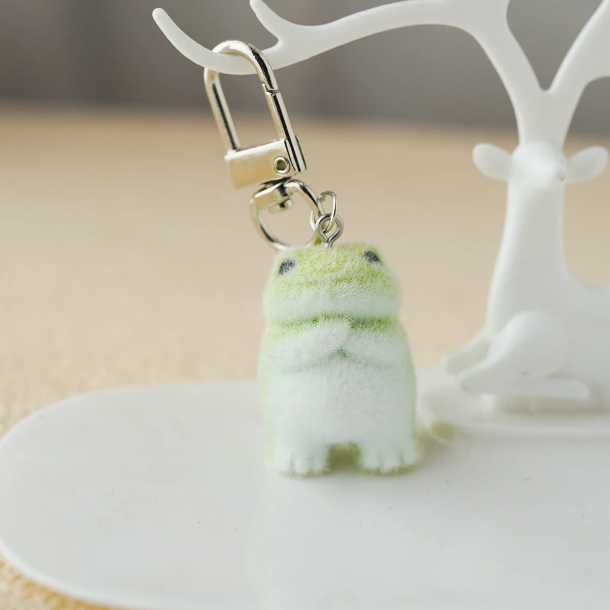 A tiny plush emotional‑support frog keychain with soft green fuzz, dangling from a metal clasp on a white stand like a little guy awaiting adoption